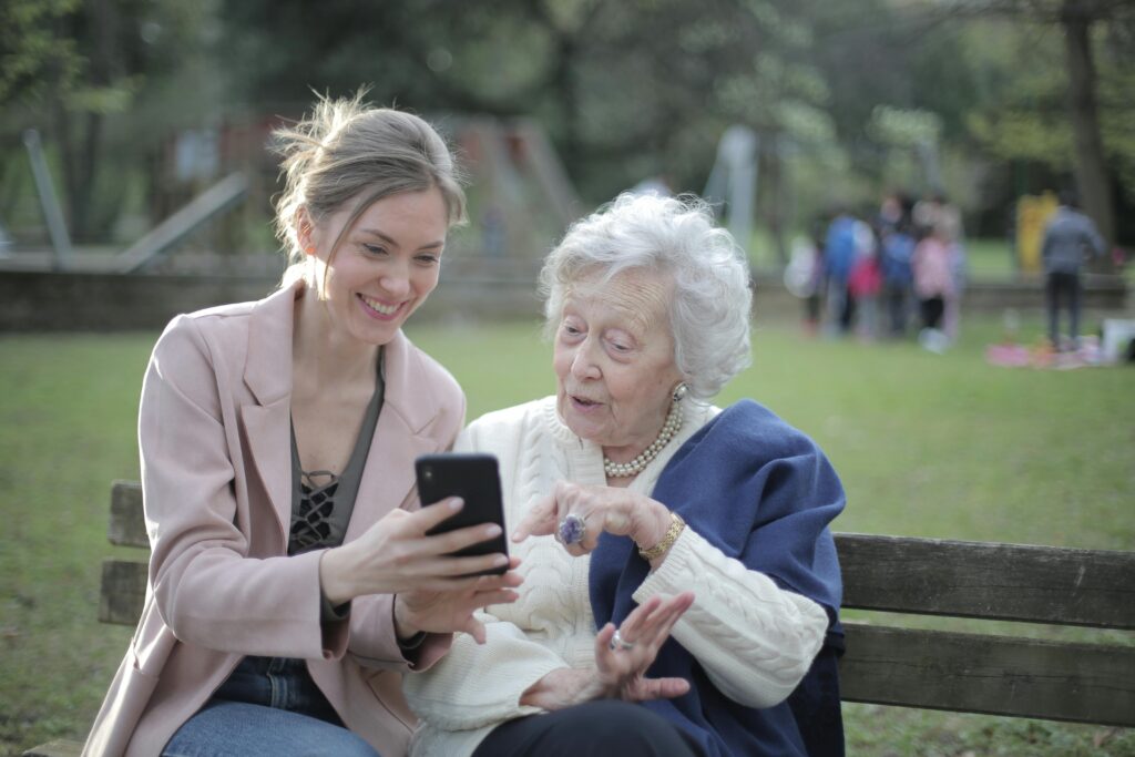 pexels-photo-3791664-3791664 Delighted female relatives sitting together on wooden bench in park and browsing mobile phone while learning using